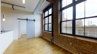 This single-floor, two-bedroom, one-bathroom residence showcases a compelling blend of industrial and modern design elements. The living area is defined by original exposed brick walls, which serve as a striking architectural backdrop and lend the space an authentic, loft-inspired character. Large, black-framed multi-pane windows dominate the brick exterior wall, flooding the interior with abundant natural light while offering views of the surrounding urban streetscape. The warmth of the natural hardwood floors throughout provides a welcoming contrast to the raw brick and industrial accents. A distinctive barn-style sliding door adds a functional yet stylish touch, contributing to the open, flexible feel of the layout. Overhead, an exposed cylindrical ductwork element runs along the ceiling, reinforcing the industrial aesthetic while maintaining the clean white upper walls. The combination of high ceilings, expansive windows, and thoughtfully preserved original building details creates a dynamic and visually engaging living environment. The two bedrooms offer comfortable accommodations, while the single bathroom completes this well-appointed residence suited for those who appreciate urban industrial design.