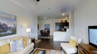 This single-floor residence showcases an open-concept layout that seamlessly connects the living, dining, and kitchen areas into one cohesive living space. Dark hardwood flooring runs throughout, providing a rich contrast against the neutral white walls and light ceilings equipped with recessed lighting.

The living area offers generous room for seating arrangements, with a floor lamp adding warm ambient light. A decorative area rug anchors the space, complementing the hardwood floors beneath. The room flows naturally into a dedicated dining area, which comfortably accommodates a round table and chairs, accented by a landscape painting on the wall.

The kitchen features a peninsula-style counter with a light-toned surface, providing both workspace and a visual divider between the kitchen and dining zone. Dark cabinetry contrasts the lighter countertops, and black appliances including a refrigerator complete the culinary space. A window above the sink allows for natural light.

The entry door is positioned conveniently off the living area, and the overall layout is well-organized, functional, and designed to maximize the sense of openness across the single floor.