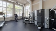 This fitness center is thoughtfully designed to support a comprehensive workout experience within an architecturally refined space. The room features soaring ceilings with decorative crown molding and elegant arched detailing, lending a sense of openness and sophistication rarely found in a gym setting. Ceiling fans provide comfortable air circulation throughout.

Large plantation-shuttered windows line multiple walls, flooding the interior with natural light and offering views of lush tropical landscaping and an outdoor pool area. Glass block wall panels and white-framed glass doors further enhance the bright, airy atmosphere.

The flooring throughout is durable dark rubber gym matting, ideal for heavy equipment and high-impact activities. The space is outfitted with a diverse selection of fitness equipment, including a treadmill, elliptical trainer, rowing machine, multi-station cable and weight machine, and an adjustable workout bench. A full rack of rubber hex dumbbells in graduated weights provides ample options for strength training, while a medicine ball rack adds versatility for functional fitness routines. A BOSU balance trainer rounds out the available equipment.

The warm neutral wall tones combined with the crisp white architectural trim create a clean, motivating environment. Overall, this fitness center balances practical functionality with an upscale aesthetic.