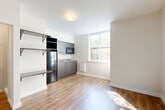Located at 3635 Phinney Ave N in Seattle's vibrant Phinney Ridge neighborhood, this studio unit offers a thoughtfully designed single-floor layout that efficiently combines living, dining, and kitchen functions into one open space. Warm-toned luxury vinyl plank flooring runs throughout, creating a cohesive and easy-to-maintain foundation. A large window floods the living area with natural light and frames views of surrounding greenery, lending an airy feel to the compact footprint. The kitchenette is outfitted with dark-toned cabinetry, a stainless steel refrigerator, an under-cabinet sink, a built-in microwave, and a wall-mounted baseboard heater nearby. Open wall shelving adjacent to the kitchen provides additional storage and display space. The main living area features clean white walls and a neutral palette, offering a versatile canvas for various furniture arrangements. An electrical panel is conveniently integrated near the entry door. The bathroom features large-format white subway tile in the shower/tub surround, complete with a recessed niche for toiletries and a glass door enclosure, presenting a clean and modern aesthetic. Overall, the unit delivers a minimalist, contemporary design well-suited for efficient urban living.