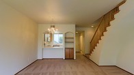 This two-story home offers a well-connected layout anchored by an open main living area finished with neutral carpet and light-toned walls that create a clean, welcoming atmosphere. The living space flows naturally toward a pass-through kitchen counter, which features a window that brings in natural light and overlooks the adjacent area. The kitchen includes warm wood cabinetry beneath the counter, providing functional storage while maintaining visual continuity with the rest of the main floor.

An elegant chandelier serves as a focal point overhead, adding a decorative touch to the dining or living zone. A graceful arched opening frames a built-in shelving unit recessed into the wall, offering both display and storage options while contributing architectural character to the space. Recessed lighting supplements the natural light throughout the main level.

A staircase with light wood railings and balusters rises along one wall, offering access to the second floor and adding a structural design element to the interior. The overall aesthetic of the main floor is cohesive and neutral, with thoughtful architectural details that balance practicality and style across both levels of the home.