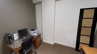 This single-floor office space offers a practical and functional environment designed for individual or administrative use. The room features neutral, cream-colored walls paired with a low-pile, beige-toned carpet that contributes to a quiet, professional atmosphere. A built-in or freestanding workstation with a granite-style countertop is positioned along one wall, accompanied by a wooden cabinet below for storage. The desk area accommodates a desktop computer monitor, keyboard, and a compact laser printer, making it well-suited for day-to-day office tasks. A standard ergonomic office chair completes the workstation setup. The walls are clean and largely unadorned, offering flexibility for personalization or the addition of shelving and equipment. Recessed structural elements along the ceiling add subtle architectural interest to the otherwise straightforward layout. A standard electrical outlet is visible on the main wall, and the space benefits from overhead lighting. Overall, the office presents a no-frills, efficient workspace well-suited for administrative, clerical, or private professional use.