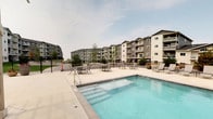 This outdoor amenity space is centered around a generously sized swimming pool with clear blue water, accessible via built-in entry steps on one end. The pool deck is finished in smooth concrete, providing ample space for lounging and circulation. A row of sling-style lounge chairs lines the poolside, offering dedicated sunbathing areas, while additional patio tables and chairs are positioned throughout the deck for casual outdoor seating.

A wood-framed pergola anchors one end of the patio area, providing a shaded gathering spot and adding architectural interest to the space. Decorative planters with seasonal plantings are placed throughout the courtyard, contributing greenery and visual warmth to the hardscaped environment.

The pool area is enclosed by a black iron perimeter fence, providing security while maintaining open sightlines. The surrounding multi-story residential buildings, finished in a contemporary combination of dark gray siding, light stone accents, and white trim, frame the courtyard on multiple sides. Each building features private balconies that overlook the shared amenity space. The open-air setting benefits from natural light throughout the day, creating a welcoming and functional outdoor retreat.