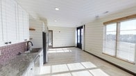 This single-floor space offers a clean, modern layout designed to maximize openness and natural light. The interior features light-toned luxury vinyl flooring that runs throughout, contributing to a cohesive and contemporary feel. Recessed lighting in the smooth white ceiling provides consistent illumination, complementing the abundant daylight that streams in through multiple windows fitted with blinds and a bold black-framed glass-panel exterior door.

The kitchen is outfitted with white cabinetry, stone-look countertops, a stainless steel sink, and a stainless steel refrigerator, offering a functional and stylish workspace. Open shelving adds a practical, modern touch. The kitchen flows seamlessly into the open living area, creating a spacious, flexible environment well-suited for a variety of uses.

The walls carry a subtle horizontal texture in a neutral cream tone, adding visual interest without overwhelming the space. Dark wood baseboards provide a grounding contrast to the lighter walls and floors. A doorway visible from the main area suggests additional rooms beyond, hinting at further functional space. Overall, the unit presents a fresh, well-lit, and move-in-ready environment with thoughtful modern finishes throughout.