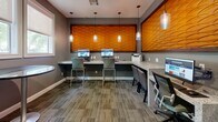 Located in Memphis, TN, this business center is a thoughtfully designed, single-room workspace that balances professional functionality with contemporary style. The space is finished with neutral gray walls, white baseboards, and patterned carpet tiles that lend a polished, corporate feel throughout.

Upon entry, a welcoming foyer greets visitors with a rich, dark wood-framed glass door, a cushioned side chair, and an abstract framed artwork that adds a touch of personality. Natural light filters in through double windows dressed with roller shades, offering views of surrounding greenery.

The main work area is the centerpiece of the space, featuring an L-shaped granite-topped workstation lined with multiple computer monitors and a printer, accommodating several users simultaneously. Overhead, a striking textured wall panel in warm amber tones commands attention, framed like a piece of art and illuminated by elegant glass pendant lights suspended from the ceiling. Additional seating along the counter includes both ergonomic task chairs and sleek bar-height stools, creating flexible work configurations.

A small bistro table with a round glass top and a guest chair complete the space, offering a casual spot for brief meetings or individual work. The overall ambiance is professional yet inviting, making this business center a well-equipped and visually appealing environment for focused work.