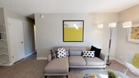 This single-storey space presents a thoughtfully designed living area that balances modern aesthetics with comfortable functionality. The living room serves as the focal point, anchored by a sleek grey sectional sofa dressed with a mix of monochromatic and patterned throw pillows in black, white, and grey. A bold abstract artwork featuring a striking yellow-and-neutral composition is mounted on the soft grey wall, introducing a vibrant accent that enlivens the otherwise understated colour scheme. A contemporary arc floor lamp positioned to one side provides both ambient and task lighting, complementing the room's clean, modern sensibility. The flooring throughout is carpeted in a warm neutral tone, adding softness underfoot and contributing to the room's relaxed atmosphere. A glass-topped coffee table is partially visible in the foreground, reinforcing the contemporary design direction. The textured ceiling and smooth walls frame the space neatly, while a hallway visible in the background hints at further rooms beyond. Overall, the space conveys a cohesive, well-curated modern interior with a calm, neutral foundation punctuated by carefully chosen decorative elements.