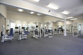 This fitness center is a comprehensively equipped, single-floor facility designed to support a wide range of workout routines. The space is generously proportioned, offering an open and airy atmosphere enhanced by high ceilings, crown molding, and an abundance of natural light streaming through numerous large windows that frame views of surrounding greenery.

The facility is organized into distinct functional zones. The cardio area features multiple treadmills and an elliptical trainer, positioned near windows to allow exercisers to enjoy outdoor views during their workouts. A wall-mounted television provides additional entertainment. The strength training section is well-appointed with a variety of cable machines, weight benches, and barbell racks, while full-length mirrors along one wall allow users to monitor their form. A dedicated free weights area houses a complete rack of dumbbells alongside stability balls and exercise mats for stretching and floor work.

Ceiling fans throughout the space promote air circulation, complementing the bright fluorescent lighting overhead. The flooring consists of durable, low-pile carpet fitted with protective rubber mats beneath equipment. A built-in water fountain and a restroom provide added convenience. The neutral color palette of whites and warm creams, combined with white structural columns, lends the facility a clean, welcoming, and professional ambiance.