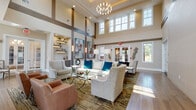 Located at 1 John F Kennedy Blvd in Somerset, NJ, this single-floor clubhouse amenity space is designed to impress with a sophisticated blend of modern and transitional design elements. The grand main lounge commands attention with its soaring double-height ceilings, an oversized multi-tiered chandelier, and large clerestory windows that flood the space with natural light. Elegant seating arrangements featuring patterned wingback chairs, a plush white sofa with teal accent pillows, and a glass-top coffee table create an inviting atmosphere, complemented by built-in display shelving and bold large-scale artwork on the walls. A striking stacked-stone accent wall anchors a sleek linear fireplace, adding warmth and texture to the adjacent seating area furnished with rich leather chairs. Wide-plank hardwood flooring flows throughout, unifying the various gathering zones. A sunlit sitting nook with floor-to-ceiling windows offers direct views of the outdoor pool area, providing a bright and serene retreat. The entertainment and game room features a billiards table, wall-mounted flat-screen TVs set against a wood-paneled accent wall, a bar area, and a dining counter with barstool seating. Additional lounge alcoves with stacked-stone accent walls and globe pendant lighting offer more intimate settings for socializing.