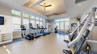 Located at 116 Danbury Rd in Wilton, CT, this fitness center offers a thoughtfully designed single-floor exercise space that balances functionality with an inviting architectural aesthetic. The room features elegant tray ceilings with ceiling fans, providing both visual height and comfortable air circulation throughout the space. Large windows along multiple walls flood the interior with natural light, creating an energizing and open atmosphere.

The fitness floor is finished with durable, speckled rubber-composite flooring suited for high-traffic exercise use. Cardio equipment includes multiple treadmills and elliptical trainers, each equipped with individual display screens, alongside a stationary recumbent cycle. A dedicated strength training section houses a comprehensive selection of selectorized cable and resistance machines, as well as a full dumbbell rack with mirrors for form monitoring.

A wall-mounted flat-screen television is positioned for easy viewing from the cardio area, while built-in open shelving provides organized storage for accessories and personal items. French-style glass-panel doors connect to an adjacent area, enhancing the sense of openness. Recessed lighting supplements natural daylight for consistent illumination. The overall ambiance is clean, bright, and well-organized, making this fitness center a practical and pleasant environment for a wide range of workout routines.
