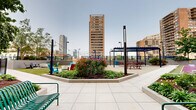 Located at 123 Montgomery St in Jersey City, NJ, this expansive rooftop amenity deck offers a diverse array of outdoor spaces designed for relaxation, recreation, and socializing, all set against a dramatic urban skyline backdrop. The deck is thoughtfully organized into distinct zones, each serving a unique purpose. A sleek outdoor pool takes center stage in one section, surrounded by a textured deck surface and framed by lush greenery and a brick accent wall, with towering Jersey City high-rises providing a striking backdrop. Adjacent to the pool, a dedicated games area features a shuffleboard court and cornhole, set on colorful turf surfaces. A well-equipped outdoor kitchen with multiple built-in grills and a pergola-covered dining area with picnic-style seating offers an ideal space for gatherings. Two fire pit lounges, each surrounded by colorful Adirondack chairs and stone accents set into lush artificial turf, provide cozy spots for evening relaxation. A vibrant children's playground, complete with climbing structures and soft turf, adds family-friendly appeal. Decorative perforated fencing lines the perimeter, while string lights, raised planters with seasonal blooms, and mature trees enhance the overall ambiance of this urban outdoor retreat.