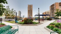 Located at 123 Montgomery St in Jersey City, NJ, this expansive rooftop amenity deck offers a diverse array of outdoor spaces designed for relaxation, recreation, and socializing, all set against a dramatic urban skyline backdrop. The deck is thoughtfully organized into distinct zones, each serving a unique purpose. A sleek outdoor pool takes center stage in one section, surrounded by a textured deck surface and framed by lush greenery and a brick accent wall, with towering Jersey City high-rises providing a striking backdrop. Adjacent to the pool, a dedicated games area features a shuffleboard court and cornhole, set on colorful turf surfaces. A well-equipped outdoor kitchen with multiple built-in grills and a pergola-covered dining area with picnic-style seating offers an ideal space for gatherings. Two fire pit lounges, each surrounded by colorful Adirondack chairs and stone accents set into lush artificial turf, provide cozy spots for evening relaxation. A vibrant children's playground, complete with climbing structures and soft turf, adds family-friendly appeal. Decorative perforated fencing lines the perimeter, while string lights, raised planters with seasonal blooms, and mature trees enhance the overall ambiance of this urban outdoor retreat.