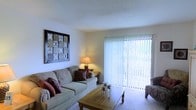 Located at 4051 E Olive Rd in Pensacola, Florida, this single-story residence offers a comfortable and well-appointed living environment. The living room serves as the heart of the home, featuring light-toned carpeting and white walls that create a clean, airy atmosphere. A large sliding glass door fitted with vertical blinds allows abundant natural light to fill the room while providing direct access to the outdoors and a view of the surrounding greenery. The textured ceiling adds a classic touch to the space. The living area comfortably accommodates a full sofa arrangement centered around a wooden coffee table, making it well-suited for both relaxing and entertaining. Warm accent lighting from a table lamp complements the natural light, contributing to an inviting ambiance throughout the day and evening. The overall design of the home emphasizes functionality and comfort, with a straightforward, single-level layout that ensures easy accessibility throughout. Situated in Pensacola, FL, the property benefits from a established neighborhood setting, offering convenient access to local amenities while maintaining a quiet residential feel.