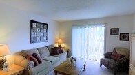 Located at 4051 E Olive Rd in Pensacola, Florida, this single-story residence offers a comfortable and well-appointed living environment. The living room serves as the heart of the home, featuring light-toned carpeting and white walls that create a clean, airy atmosphere. A large sliding glass door fitted with vertical blinds allows abundant natural light to fill the room while providing direct access to the outdoors and a view of the surrounding greenery. The textured ceiling adds a classic touch to the space. The living area comfortably accommodates a full sofa arrangement centered around a wooden coffee table, making it well-suited for both relaxing and entertaining. Warm accent lighting from a table lamp complements the natural light, contributing to an inviting ambiance throughout the day and evening. The overall design of the home emphasizes functionality and comfort, with a straightforward, single-level layout that ensures easy accessibility throughout. Situated in Pensacola, FL, the property benefits from a established neighborhood setting, offering convenient access to local amenities while maintaining a quiet residential feel.
