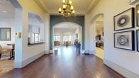 This generously sized single-floor space showcases a thoughtfully designed interior that blends elegance with functionality. The grand entryway immediately impresses with its soaring ceilings, rich dark hardwood flooring, and a striking ornate chandelier suspended beneath a coffered ceiling. Dramatic arched doorways framed by teal accent walls draw the eye through the space, creating a seamless flow between areas.

The open-plan living area is bathed in natural light from large windows dressed with elegant draping curtains, and features a substantial sectional sofa set atop a patterned area rug. Adjacent to this is a welcoming dining area that benefits from the same abundance of natural light.

The gourmet kitchen is a standout feature, offering rich dark espresso cabinetry, granite countertops with under-cabinet lighting, decorative tile backsplash with artisan detailing, and stainless steel appliances. A curved breakfast bar with pendant lighting provides additional seating and serves as a natural gathering point.

A dedicated office area presents a professional setting with patterned carpet, dual workstations, accent chairs, and a modern geometric pendant light. Multiple windows allow generous natural light and outdoor views. The overall ambiance of the space is refined yet inviting, combining warm tones with architectural character throughout.