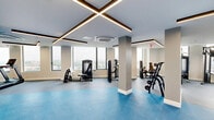 Located on an upper floor at 626 Summit Ave in Jersey City, NJ, this expansive fitness center offers a well-appointed exercise environment designed for comprehensive training. The space is characterized by a clean, modern aesthetic, featuring neutral-toned walls, striking geometric LED linear lighting accents on the ceiling, and a combination of rubberized gray and vibrant blue flooring that visually delineates distinct workout zones.

Large floor-to-ceiling windows line the perimeter, flooding the interior with abundant natural light and offering sweeping elevated views of the Jersey City skyline and surrounding neighborhoods — an energizing backdrop for any workout. Ceiling-mounted HVAC cassette units ensure consistent climate control throughout the space.

The cardio zone is well-equipped with a row of treadmills, elliptical trainers, and stair climbers positioned along the windowed wall to maximize the outdoor views. The strength training area features an extensive dumbbell rack with a full weight selection, adjustable benches, cable and weight machines, and a functional training rig complete with pull-up bars and battle rope attachments. Full-length mirrors throughout enhance spatial depth and allow users to monitor form. The facility's generous footprint provides ample open floor space, supporting a wide variety of training styles in a bright, modern, and motivating environment.