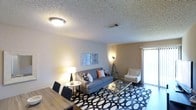 This single-floor space offers a comfortable and stylishly designed interior that balances modern aesthetics with functional living. The living area serves as the focal point of the space, featuring a plush gray sofa accented with decorative pillows, a sleek glass-top coffee table, and a classic white accent chair. A bold geometric-patterned area rug anchors the seating arrangement, adding a contemporary touch to the room. Warm ambient lighting is provided by a combination of a flush-mount ceiling fixture and complementary floor and table lamps, creating an inviting atmosphere throughout. The walls are adorned with modern artwork, contributing to the overall curated feel of the space. A sliding glass door fitted with vertical blinds allows natural light to filter in while offering privacy, and hints at outdoor access. Adjacent to the living area, a dining space with dark chairs and a wood-toned table provides a practical area for meals or work. The textured ceiling adds architectural character to the interior. Overall, the space presents a cohesive, move-in-ready environment with thoughtful attention to design detail and everyday functionality.