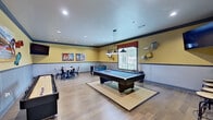 This single-floor recreational facility is designed for entertainment and social interaction, offering a well-appointed game room alongside an elegant lounge corridor. The game room is the centerpiece of the space, featuring light hardwood flooring throughout and warm yellow walls accented by a two-tone wainscoting detail with a bold blue chair rail. A full-size pool table anchors the room beneath a trio of industrial-style pendant lights, with natural light streaming in through large windows fitted with blinds. Surrounding the pool table are additional game options, including a shuffleboard table, a foosball table, and a checkers-top dining table with modern industrial chairs. Colorful artwork and eclectic wall-mounted decorative pieces add personality throughout, while a wall-mounted flat-screen TV provides additional entertainment. A high counter with bar-height stools offers a spot to watch the action. The adjacent lounge corridor transitions the space into a more refined setting, with crisp white walls, recessed lighting, and large-scale artwork including a striking equine painting. A natural stone accent wall frames a wood-mantel fireplace topped with a mounted TV, creating a warm and inviting gathering area.
