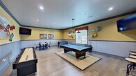 This single-floor recreational facility is designed for entertainment and social interaction, offering a well-appointed game room alongside an elegant lounge corridor. The game room is the centerpiece of the space, featuring light hardwood flooring throughout and warm yellow walls accented by a two-tone wainscoting detail with a bold blue chair rail. A full-size pool table anchors the room beneath a trio of industrial-style pendant lights, with natural light streaming in through large windows fitted with blinds. Surrounding the pool table are additional game options, including a shuffleboard table, a foosball table, and a checkers-top dining table with modern industrial chairs. Colorful artwork and eclectic wall-mounted decorative pieces add personality throughout, while a wall-mounted flat-screen TV provides additional entertainment. A high counter with bar-height stools offers a spot to watch the action. The adjacent lounge corridor transitions the space into a more refined setting, with crisp white walls, recessed lighting, and large-scale artwork including a striking equine painting. A natural stone accent wall frames a wood-mantel fireplace topped with a mounted TV, creating a warm and inviting gathering area.