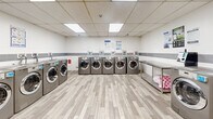 Located at 2801 Jane St in Toronto, Ontario, this well-appointed commercial laundry facility offers a spacious, single-floor layout designed for efficient, high-volume use. The room is bright and clean, illuminated by recessed fluorescent panel lighting set into a drop-tile ceiling that keeps the space feeling open and well-lit throughout.

The facility is equipped with a substantial row of front-load washing machines and dryers finished in a sleek stainless steel tone, arranged along the perimeter walls to maximize the usable floor space. Stainless steel folding and sorting tables are conveniently positioned throughout the room, providing ample surface area for laundry preparation. A freestanding deep utility sink is centrally located along one wall, offering a practical handwashing and pre-treatment station.

The walls feature a clean white finish accented by a horizontal band of grey tile, contributing to a tidy, hygienic aesthetic. The floor is covered in wood-look vinyl plank tiles, adding a touch of warmth while remaining easy to maintain. A stainless steel waste bin and bulletin board with posted instructions and notices round out the functional amenities. Access to the facility is provided through secure grey doors. The overall environment is clean, organized, and purpose-built to serve a busy residential community.