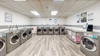 Located at 2801 Jane St in Toronto, Ontario, this well-appointed commercial laundry facility offers a spacious, single-floor layout designed for efficient, high-volume use. The room is bright and clean, illuminated by recessed fluorescent panel lighting set into a drop-tile ceiling that keeps the space feeling open and well-lit throughout.

The facility is equipped with a substantial row of front-load washing machines and dryers finished in a sleek stainless steel tone, arranged along the perimeter walls to maximize the usable floor space. Stainless steel folding and sorting tables are conveniently positioned throughout the room, providing ample surface area for laundry preparation. A freestanding deep utility sink is centrally located along one wall, offering a practical handwashing and pre-treatment station.

The walls feature a clean white finish accented by a horizontal band of grey tile, contributing to a tidy, hygienic aesthetic. The floor is covered in wood-look vinyl plank tiles, adding a touch of warmth while remaining easy to maintain. A stainless steel waste bin and bulletin board with posted instructions and notices round out the functional amenities. Access to the facility is provided through secure grey doors. The overall environment is clean, organized, and purpose-built to serve a busy residential community.