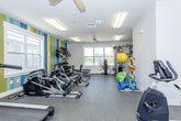 This fitness center is a thoughtfully designed, single-floor space offering a comprehensive range of exercise equipment and amenities. The room is immediately distinguished by a bold, vertically striped accent wall in shades of teal, blue, yellow, and white, lending a vibrant and energizing atmosphere to the space.

Natural light filters in through multiple windows positioned along the walls, providing views of the surrounding exterior while keeping the environment bright and welcoming. Overhead, a ceiling fan complements the flush-mounted fluorescent light panels, ensuring consistent air circulation and illumination throughout.

The main workout area is well-organized, housing a row of Matrix-brand cardio machines that includes ellipticals, treadmills, and upright stationary bikes. A dedicated section along the accent wall features a wall-mounted flat-screen television, a multi-tiered dumbbell rack stocked with a full range of free weights, and an adjustable weight bench, catering to strength training needs. A cable resistance machine is also available for functional training exercises.

A separate, quieter alcove connects to the main room and accommodates two recumbent stationary bikes, offering a lower-impact cardio option. Stability balls, medicine balls, and yoga mats are stored along one wall, supporting stretching and functional fitness routines. The durable carpet-style flooring throughout provides cushioning and traction suitable for varied exercise activities.