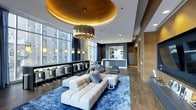 This single-floor space exemplifies contemporary design with a refined, upscale aesthetic. The centerpiece of the room is a dramatic circular gold ceiling medallion with recessed lighting, from which a cylindrical brass chandelier descends, casting a warm, ambient glow throughout the space. The open-plan layout is generous and well-appointed, seamlessly integrating multiple functional zones.

A plush, low-profile sectional sofa in cream anchors the lounge area, positioned atop a deep blue area rug that adds a rich contrast to the warm wood flooring. Accent chairs in charcoal provide additional seating, and a sleek side table completes the arrangement.

Along the perimeter, floor-to-ceiling windows allow abundant natural light and offer views of the surrounding urban skyline. A row of counter-height bar seating lines the window wall, illuminated by pendant lights above, creating an ideal setting for socializing or working.

Toward the rear, a service counter or reception area is framed by wood-paneled walls and a built-in display niche, adding depth and warmth. The overall ambiance is polished and cosmopolitan, blending modern architecture with luxurious finishes.