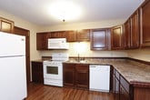This single-floor space is thoughtfully laid out across several distinct areas, each finished with consistent design elements that give the unit a cohesive feel. The living areas are covered in neutral-toned carpet and feature ceiling fans with light fixtures, textured ceilings, and a notable two-tone wall treatment — warm beige upper walls paired with a darker wainscoting below, separated by a decorative border trim. Sliding glass doors in the main living space allow natural light to fill the room and provide views of surrounding greenery.

The kitchen is well-equipped, featuring rich dark-stained wood cabinetry that extends to the ceiling, granite-look laminate countertops, and wood-look plank flooring. White appliances — including a range, built-in microwave, dishwasher, and refrigerator — are included. A front entry area adjacent to the kitchen provides a practical transition space.

The bathroom includes a dark wood vanity with an integrated sink, stone-look countertop, a mirrored medicine cabinet, and a tub-shower combination with dark fixtures. The dedicated laundry room offers additional dark cabinetry with matching countertop, a clothing rod for hanging, and space for a full-size washer and dryer.