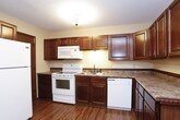 This single-floor space is thoughtfully laid out across several distinct areas, each finished with consistent design elements that give the unit a cohesive feel. The living areas are covered in neutral-toned carpet and feature ceiling fans with light fixtures, textured ceilings, and a notable two-tone wall treatment — warm beige upper walls paired with a darker wainscoting below, separated by a decorative border trim. Sliding glass doors in the main living space allow natural light to fill the room and provide views of surrounding greenery.

The kitchen is well-equipped, featuring rich dark-stained wood cabinetry that extends to the ceiling, granite-look laminate countertops, and wood-look plank flooring. White appliances — including a range, built-in microwave, dishwasher, and refrigerator — are included. A front entry area adjacent to the kitchen provides a practical transition space.

The bathroom includes a dark wood vanity with an integrated sink, stone-look countertop, a mirrored medicine cabinet, and a tub-shower combination with dark fixtures. The dedicated laundry room offers additional dark cabinetry with matching countertop, a clothing rod for hanging, and space for a full-size washer and dryer.
