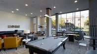This contemporary single-floor amenity space is designed to offer a dynamic and social environment with multiple activity zones seamlessly integrated into one open layout. At the center of the room sits a full-sized pool table illuminated by a sleek linear pendant light, serving as the focal point of the space. The room benefits from abundant natural light streaming through large floor-to-ceiling windows that frame views of the surrounding outdoor landscape, lending the interior an open and airy feel.

The lounge area is furnished with comfortable sofas and accent tables, providing a relaxed setting for casual gatherings. A dining or meeting zone features chairs arranged around tables, accompanied by a wall-mounted television, making it suitable for both social and functional use. Additional game seating and a standing-height game table can be spotted near the windows, enhancing the recreational offerings of the space.

Architectural details include recessed ceiling lighting, square structural columns with clean, neutral finishes, and neutral-toned carpeting throughout. The overall ambiance is modern and inviting, balancing leisure and functionality in a well-lit, thoughtfully designed communal environment.