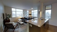 This contemporary single-floor residence showcases a thoughtfully designed open-concept layout that unifies the kitchen, dining, and living areas into one cohesive, light-filled space. Large corner windows spanning nearly the full width of two walls flood the interior with natural light and offer sweeping views of the surrounding neighborhood and skyline, creating an airy, connected atmosphere throughout.

The kitchen is anchored by a substantial island with a warm, neutral countertop finish and under-counter accent lighting, providing both a functional workspace and a natural gathering point. A stainless steel sink and pendant lighting above the island add refined, practical detail to the space.

The open living area features warm hardwood flooring that flows seamlessly throughout, grounding the space with texture and warmth. The living zone comfortably accommodates a sectional sofa, accent chairs, and a central coffee table, all layered over a patterned area rug. A dedicated dining area sits adjacent, benefiting from the same generous window exposure.

The overall aesthetic is clean and modern, with a neutral white palette on the walls and ceilings complemented by warm wood tones and contemporary fixtures. The combination of abundant natural light, open sightlines, and cohesive design details makes this a bright, functional, and welcoming living space.