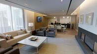 This refined single-floor space presents a seamless open-plan design that integrates a living area, dining space, and kitchen into one cohesive, flowing environment. The living area is anchored by a sleek marble-topped coffee table and a pair of contemporary grey upholstered armchairs, complemented by a neutral sofa adorned with accent pillows and a stylish table lamp with a chrome base. A wall-mounted flat-screen television is set against a warm gold-toned accent wall, adding depth and visual contrast to the space. The flooring transitions from a patterned area rug in the lounge zone to light hardwood in the dining and kitchen areas, creating a subtle yet effective delineation between spaces. The dining area features a clean-lined table with modern dark chairs, positioned adjacent to the kitchen, which showcases marble-clad walls and contemporary pendant lighting. Recessed ceiling lighting throughout provides a polished, well-illuminated ambiance, while large windows allow natural light to filter in, enhancing the bright and airy atmosphere. The overall aesthetic is modern and sophisticated, with a carefully curated neutral palette punctuated by rich textures and refined finishes.