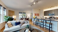 This thoughtfully designed single-floor residence offers a seamless open-concept layout that connects the living, dining, and kitchen areas into one cohesive, light-filled space. Warm hardwood-style flooring flows throughout the main living areas, complemented by neutral walls and contemporary track lighting overhead.

The living room is bright and inviting, anchored by large windows fitted with roller shades that frame outdoor views. The kitchen is well-appointed with slate-gray cabinetry, stainless steel appliances including a gas range, microwave, and refrigerator, granite-style countertops, and a classic white subway tile backsplash. A breakfast bar with seating connects the kitchen to the main living space. The dedicated dining area features generous window exposure with views of outdoor greenery, creating a pleasant setting for meals.

The primary bedroom is a comfortable retreat with plush carpeting, ample closet space, and natural light from a large window. It is served by a private en-suite half bathroom. A second full bathroom includes a tub-shower combination, floating wood vanity, and clean tiled finishes. The second bedroom is equally well-lit with bright window exposure and carpeted flooring. A dedicated laundry area with a full-size washer provides added convenience. The entryway is welcoming, with hardwood-style flooring and a paneled front door with a smart lock.