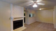 This single-story space centers around a living area with a striking white-painted fireplace as its focal point. The fireplace features a bold diagonal wood-panel surround extending to the ceiling, a classic mantel, and a tiled hearth, adding both warmth and visual interest to the room. Neutral-toned carpeting runs throughout, contributing to a cozy, cohesive feel. A ceiling fan with an integrated light fixture is mounted overhead, providing both comfort and ambient lighting. The walls are finished in a soft, warm neutral tone that complements the overall palette. A notable architectural detail is the pass-through window cut into the wall, connecting the living area to an adjacent kitchen or dining space, enhancing the open and communicative flow between rooms. The pass-through also allows natural light to travel between spaces. A standard HVAC vent is visible in the ceiling, and the layout appears straightforward and functional. The space offers a comfortable, lived-in ambiance suitable for everyday living, with its fireplace serving as an inviting gathering point in the main living area.