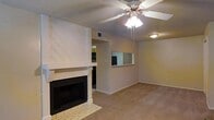 This single-story space centers around a living area with a striking white-painted fireplace as its focal point. The fireplace features a bold diagonal wood-panel surround extending to the ceiling, a classic mantel, and a tiled hearth, adding both warmth and visual interest to the room. Neutral-toned carpeting runs throughout, contributing to a cozy, cohesive feel. A ceiling fan with an integrated light fixture is mounted overhead, providing both comfort and ambient lighting. The walls are finished in a soft, warm neutral tone that complements the overall palette. A notable architectural detail is the pass-through window cut into the wall, connecting the living area to an adjacent kitchen or dining space, enhancing the open and communicative flow between rooms. The pass-through also allows natural light to travel between spaces. A standard HVAC vent is visible in the ceiling, and the layout appears straightforward and functional. The space offers a comfortable, lived-in ambiance suitable for everyday living, with its fireplace serving as an inviting gathering point in the main living area.