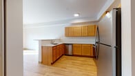 This single-floor space is thoughtfully designed with consistent warm-toned wood finishes and neutral walls throughout, creating a cohesive and functional living environment. The kitchen serves as a central feature, showcasing an L-shaped layout with honey-toned wood cabinetry extending from lower to upper sections, light-colored countertops, and stainless steel appliances including a refrigerator, range, over-the-range microwave, and dishwasher. A peninsula counter extends outward, offering additional workspace or casual dining. Light wood-style flooring runs throughout the kitchen and adjoining living area, enhancing the brightness of the space. Recessed lighting provides focused illumination overhead. The bedroom areas feature plush neutral carpeting, natural light from windows overlooking greenery, and closet access with built-in wire shelving. The bathroom continues the wood-tone theme with a matching vanity, white countertop, and large mirror above, complemented by geometric tile flooring in a soft neutral palette. The overall layout is open and practical, with a clean, move-in-ready aesthetic.