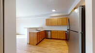 This single-floor space is thoughtfully designed with consistent warm-toned wood finishes and neutral walls throughout, creating a cohesive and functional living environment. The kitchen serves as a central feature, showcasing an L-shaped layout with honey-toned wood cabinetry extending from lower to upper sections, light-colored countertops, and stainless steel appliances including a refrigerator, range, over-the-range microwave, and dishwasher. A peninsula counter extends outward, offering additional workspace or casual dining. Light wood-style flooring runs throughout the kitchen and adjoining living area, enhancing the brightness of the space. Recessed lighting provides focused illumination overhead. The bedroom areas feature plush neutral carpeting, natural light from windows overlooking greenery, and closet access with built-in wire shelving. The bathroom continues the wood-tone theme with a matching vanity, white countertop, and large mirror above, complemented by geometric tile flooring in a soft neutral palette. The overall layout is open and practical, with a clean, move-in-ready aesthetic.