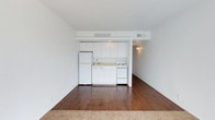 This single-floor unit presents a straightforward, open-concept layout that combines the living area and kitchen into one cohesive space. The kitchenette is neatly tucked along one wall and is equipped with white cabinetry extending to the ceiling, a white refrigerator, a white electric range, and a single-basin sink — all finished in a clean, uniform palette that contributes to a bright, orderly aesthetic. White walls throughout the space amplify the sense of openness and keep the interior feeling light. The flooring transitions from warm, rich hardwood in the main living area to carpet near the entry, adding a subtle variation in texture underfoot. A hallway visible beyond the kitchen leads to additional areas of the unit, hinting at further functional space. Overhead, HVAC vents are integrated into the ceiling, ensuring climate control throughout. The overall ambiance is clean and minimalist, making it a practical canvas well-suited for personalization.