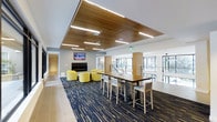 This single-floor communal space showcases a thoughtfully designed interior that balances functionality with contemporary aesthetics. The most striking architectural feature is the suspended wood-slat ceiling installation, which adds warmth and texture while housing integrated linear lighting that illuminates the space evenly throughout.

The lounge is organized into distinct zones. A seating area anchored by a sleek, wall-mounted linear fireplace is furnished with vibrant yellow armchairs, creating a relaxed and inviting atmosphere. A wall-mounted flat-screen television above the fireplace adds a media-friendly element to this lounge zone.

Adjacent to the seating area, a long communal high-top table with bar-height chairs provides a more structured setting suited for collaborative work or casual dining. The patterned carpet underfoot features a bold, multi-directional stripe design in deep navy and neutral tones, adding visual energy to the floor plane.

Floor-to-ceiling windows along one side of the space draw in abundant natural light and offer views of the surrounding greenery, enhancing the open, airy feel. The combination of tile flooring in the surrounding corridors and carpeted gathering zones helps define each area within the open-plan layout.