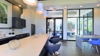 This single-floor space showcases a contemporary design aesthetic with clean lines and a carefully curated color palette of white, black, and grey. The focal point of the interior is a generous kitchen or lounge bar area anchored by a long, light-colored countertop with a contrasting dark base, accompanied by sleek, modern swivel chairs that invite casual seating.

Overhead, two large white bell-shaped pendant lights with warm copper interiors provide both functional illumination and a striking design statement. Recessed ceiling lighting supplements the overall brightness of the space.

Dark cabinetry lines one wall alongside stainless steel appliances, adding a sophisticated contrast to the lighter surfaces throughout. The dark hardwood-style flooring grounds the space and ties together the bold design elements.

A standout feature is the expansive floor-to-ceiling black-framed glass doors and windows, which flood the interior with abundant natural light and provide a seamless visual connection to an outdoor patio or courtyard area. The exterior space, visible through the glass, features comfortable seating and lush greenery, enhancing the overall sense of openness and flow throughout the space.