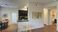 This single-floor residence offers a well-designed open-concept layout that seamlessly connects the kitchen and living areas. The kitchen is a standout feature, showcasing dark espresso cabinetry that contrasts beautifully with the dark countertops, creating a sleek and modern aesthetic. Black appliances, including a microwave and range, complement the cabinetry for a cohesive look. A peninsula-style breakfast bar with a double sink extends into the living space, providing additional seating with two bar stools — perfect for casual dining or entertaining. Rich hardwood-style flooring runs throughout the unit, adding warmth and continuity to the space. The living area benefits from natural light streaming in through a large window with blinds, lending an airy, open feel. Neutral wall tones provide a clean backdrop that allows the architectural details and finishes to take center stage. A decorative art piece adorns the wall, adding a touch of character to the space. Additional rooms, visible through open doorways, suggest a functional and thoughtfully arranged floor plan that balances comfort with style.