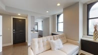 Located at 50 W Broad St in the heart of downtown Columbus, Ohio, this single-floor unit offers a thoughtfully designed open-concept layout with a contemporary aesthetic. The living area is finished with smooth neutral-toned walls, recessed ceiling lighting, and light hardwood-style flooring that extends throughout the space, creating a cohesive and inviting atmosphere.

An arched window in the living area allows natural light to filter in, offering views of the surrounding urban environment. The space transitions seamlessly into the kitchen, which features gray cabinetry, a stainless steel refrigerator, a gas range, and a light-colored countertop — providing a functional yet stylish cooking space. A pass-through or open archway connects the two areas, reinforcing the open and airy feel of the unit.

The entrance is framed by a dark-toned door that provides a subtle contrast to the otherwise light interior palette. Clean lines, recessed lighting, and a neutral color scheme throughout contribute to a modern, well-appointed feel suited for urban living in a prime downtown Columbus location.