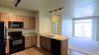 This single-floor space showcases an open-concept layout that integrates the kitchen and living area into a cohesive, functional environment. The kitchen is equipped with warm wood-toned cabinetry in two distinct finishes — lighter upper and lower cabinets along the perimeter wall paired with darker cabinetry on the peninsula island. Granite countertops with subtle gold and beige tones run throughout, providing ample prep and serving space. The peninsula island features a double stainless steel sink and a dishwasher, making it both a practical workspace and a natural divider between the kitchen and the adjoining living area. Black appliances, including a range and microwave, complement the hardware and fixtures throughout. Track lighting mounted on the ceiling directs focused illumination across the kitchen workspace. The flooring transitions from hardwood-style planks in the kitchen to plush carpet in the living area, delineating the two zones. A doorway visible from the kitchen suggests access to additional rooms beyond. The living area benefits from a large window that invites generous natural light, creating a bright and welcoming ambiance throughout the space.