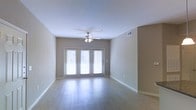 This single-floor space presents a thoughtfully designed open living area that balances simplicity with functional comfort. The room is finished with smooth light hardwood-style flooring that runs throughout, reflecting natural light and contributing to a bright, airy atmosphere. Neutral taupe-toned walls provide a versatile backdrop suitable for a variety of interior styles and furnishing choices.

A ceiling fan with an integrated light fixture is centrally mounted, offering both air circulation and ambient lighting. The room benefits significantly from a set of double French doors fitted with horizontal blinds, which serve as the primary source of natural light. These doors open outward, likely to an outdoor area, and allow daylight to flood the interior during the day.

White baseboards frame the perimeter of the room cleanly, complementing the overall neutral color palette. Ceiling-mounted HVAC vents are visible, indicating a central air system. Multiple electrical outlets are positioned at regular intervals along the walls, adding to the room's practical utility.

Overall, the space presents a clean, move-in-ready environment with a calm, understated aesthetic well-suited for comfortable everyday living.