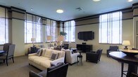 This well-appointed single-floor space presents a refined and welcoming atmosphere, thoughtfully designed for comfort and social interaction. The room features a sophisticated neutral color palette, with cream and taupe tones complemented by dark wood accents that lend a sense of elegance throughout.

The seating area is generously furnished with plush cream sofas and dark upholstered armchairs arranged around a central coffee table, creating an inviting conversational layout. A large wall-mounted flat-screen television serves as a focal point on one wall, flanked by a sleek media console, making the space well-suited for both entertainment and leisure.

An abundance of natural light pours in through multiple large windows fitted with slatted blinds, illuminating the space and offering views of the surrounding greenery. The windows are framed by a distinctive dark horizontal banding detail near the ceiling, adding architectural character to the room. Recessed lighting and stylish table lamps provide warm supplemental illumination.

The flooring is covered in a neutral, textured carpet that ties the room's design together cohesively. Decorative elements, including potted plants and accent pillows with geometric patterns, add subtle visual interest. Overall, the space exudes a polished, contemporary ambiance suited to a high-end communal lounge or hospitality setting.