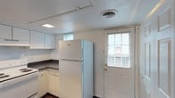 This two-floor space includes a practical kitchen area finished with white cabinetry and dark laminate countertops arranged in an L-shaped layout. The kitchen is equipped with a full-size top-freezer refrigerator and features white lower cabinets with ample storage. A skylight panel in the ceiling supplements the natural light that enters through horizontal-slatted blinds on two windows positioned at varying heights along the walls. A white paneled exterior door with a brass handle and an adjacent window provides direct outdoor access from the kitchen, adding both convenience and additional natural light to the space. The ceiling is finished with a central circular HVAC vent, contributing to efficient climate control throughout. Dark hardwood-style flooring grounds the kitchen area, providing a contrast to the light-toned walls and cabinetry. The overall design is straightforward and utilitarian, offering a clean, well-lit environment with a neutral color palette that provides a versatile foundation for personalization across both floors of the space.