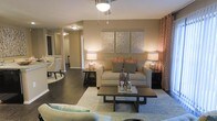This single-floor space showcases a thoughtfully designed open-concept living area that blends comfort with contemporary style. The living room serves as the heart of the space, anchored by a neutral-toned sofa accented with coral and gray throw pillows, complemented by a dark-finished rectangular coffee table and a soft area rug that defines the seating zone. Warm ambient lighting is provided by matching table lamps on either side of the sofa, along with a ceiling fan fixture overhead that adds both function and a finishing touch to the room.

The walls are painted in a warm taupe tone, creating an inviting atmosphere, while a trio of decorative wall panels above the sofa adds a refined focal point. Rich dark hardwood flooring runs throughout, offering a striking contrast to the lighter furnishings. Coral-patterned curtains introduce a pop of color and texture along the window wall.

The open layout allows for a natural visual connection to an adjacent dining area, visible in the background, enhancing the overall sense of spaciousness. The cohesive design palette and layered lighting contribute to an elegant yet approachable ambiance throughout.