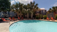 The outdoor pool area serves as a central amenity, anchored by a generously sized swimming pool with clear, turquoise water. The pool deck is finished in light-toned concrete, providing ample space for multiple lounge chairs and seating arrangements. Shaded umbrella stations are positioned along the deck, offering relief from direct sun exposure. Tall palm trees and mature landscaping surround the area, contributing a tropical ambiance and natural privacy screening. A dedicated pool house structure features textured stucco exterior walls, wood-framed entry doors, and wall-mounted lighting. Large glass sliding doors within the pool house provide a direct visual connection to the pool beyond. The surrounding multi-story building features brick and wood-toned siding with covered balconies that overlook the pool area. A separate spa or hot tub is positioned adjacent to the main pool, adding an additional amenity option. The overall setting benefits from abundant natural light and open sky views, creating an inviting outdoor retreat. The combination of lush greenery, well-maintained hardscaping, and thoughtful seating arrangements gives the space a resort-like character suited for relaxation and recreation.