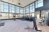 Located in Dale City, Virginia, this well-appointed fitness center delivers a bright, energizing environment designed to support a complete workout experience. The space is defined by its dramatic double-height ceilings and expansive floor-to-ceiling windows that wrap around the main exercise area, flooding the interior with natural light and offering open views of the surrounding community. The contemporary design features a bold color palette of deep charcoal and crisp white, complemented by plush, patterned carpet flooring that softens the workout area underfoot. A large full-wall mirror on one side visually expands the space and supports strength training. The main floor houses a comprehensive selection of cardio equipment, including treadmills, ellipticals, and stationary bikes, alongside a dedicated strength zone equipped with a cable machine, weight rack, and adjustable benches. Overhead, ceiling fans and pendant lighting ensure comfortable air circulation and consistent illumination. The facility includes two bathrooms for convenience, as well as a closet for storage. The overall ambiance is modern and professional, making it a fully functional and visually impressive fitness destination.