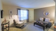 Located at 3500 Creighton Rd in Pensacola, Florida, this single-floor residential unit offers a practical and inviting layout designed for comfortable everyday living. The living area serves as the centerpiece of the home, featuring wall-to-wall carpeting that adds warmth underfoot and a soft, neutral color palette throughout. Walls are finished in a combination of warm beige and cool gray tones, creating a modern yet relaxed ambiance. A large window dressed with stylish gray grommet curtains allows ample natural light to fill the room, enhancing the open and airy feel of the space. The living area comfortably accommodates a seating arrangement, including a sofa and accent chair with decorative patterned upholstery, centered around a dark wood coffee table. A dedicated entryway door is conveniently positioned within the living space, providing direct access to the unit. The smooth ceiling and clean architectural lines throughout contribute to a tidy, well-maintained appearance. Overall, this unit presents a move-in-ready interior with a cohesive design sensibility, situated in an accessible Pensacola location close to local amenities.