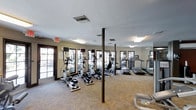 The fitness center at 3950 Leland in San Diego is a well-equipped, single-floor exercise facility designed to support a variety of cardio-focused workouts. The space is outfitted with multiple elliptical machines and treadmills arranged in a practical, open layout that allows for easy navigation and comfortable use of each station. Structural support columns are integrated throughout the floor, adding an architectural element to the space. Large, multi-pane windows line the walls, flooding the interior with abundant natural light and offering views of the surrounding greenery, creating a refreshing and motivating workout environment. Mirrored walls further enhance the sense of openness, visually expanding the room and allowing users to monitor their form during exercise. The flooring features a patterned carpet that adds warmth and texture to the space. Overhead, fluorescent lighting supplements the natural light, ensuring the facility remains bright and functional at any time of day. An emergency exit is clearly marked, reflecting attention to safety standards. The overall ambiance is clean, functional, and welcoming, making this fitness center a convenient and practical amenity for residents of the 3950 Leland community in San Diego.